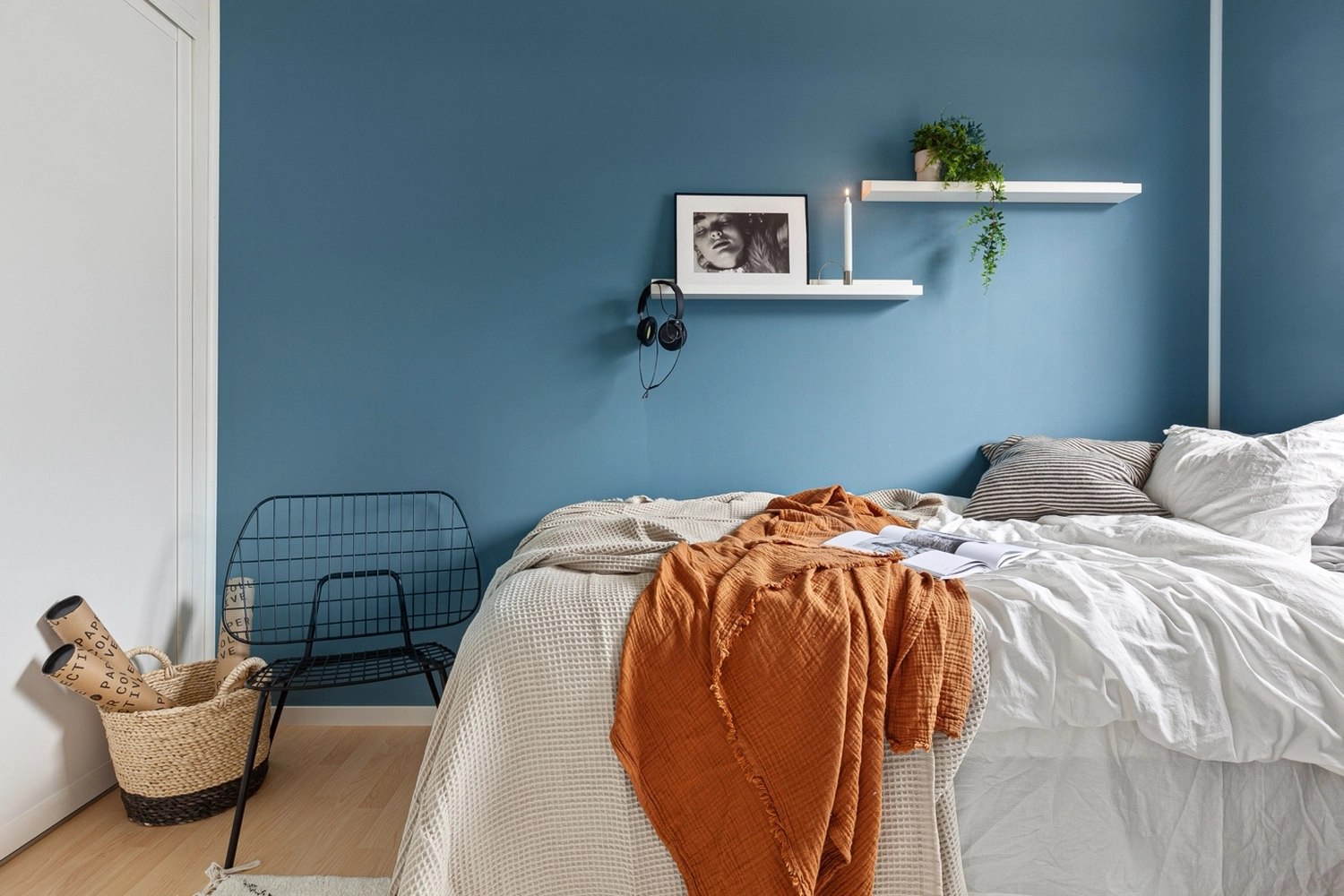 Antjes – Roomstyling – Ein gemütliches Schlafzimmer mit tiefblauen Wänden, einem Bett mit weißer und rostfarbener Bettwäsche, einem Drahtstuhl und minimalistischen Wandregalen.