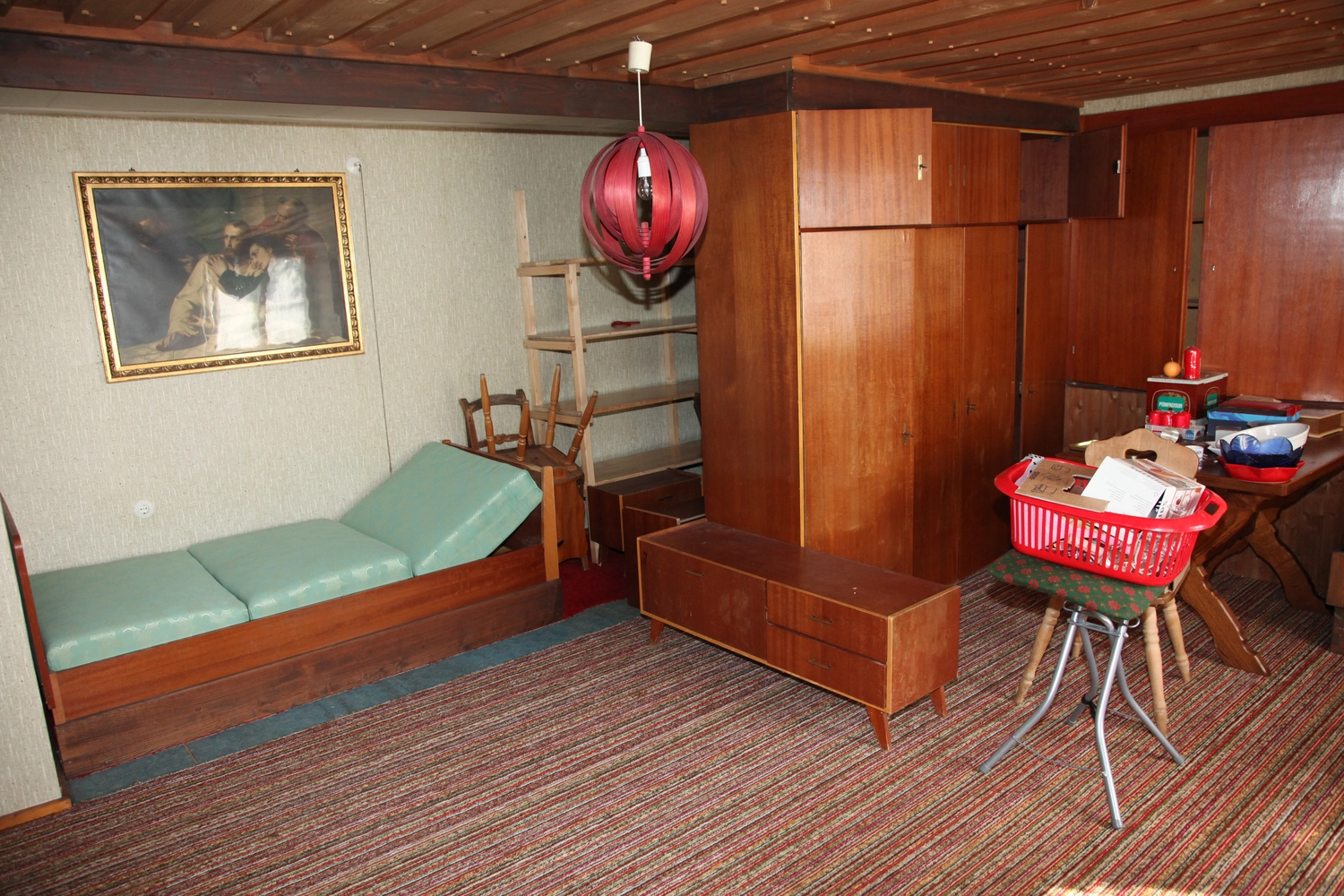 Antjes – Roomstyling –  Vorher: Ein veraltetes Wohnzimmer mit Holzmöbeln, rotem Teppich, einer grünen Liege und einer roten Hängelampe.