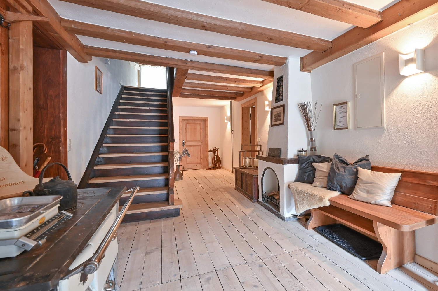 Antjes – Roomstyling –  Nachher: Ein heller Flur mit weiß gestrichenen Wänden, Holzdielen, einer modernen Treppe und einer Sitzbank mit Kissen.