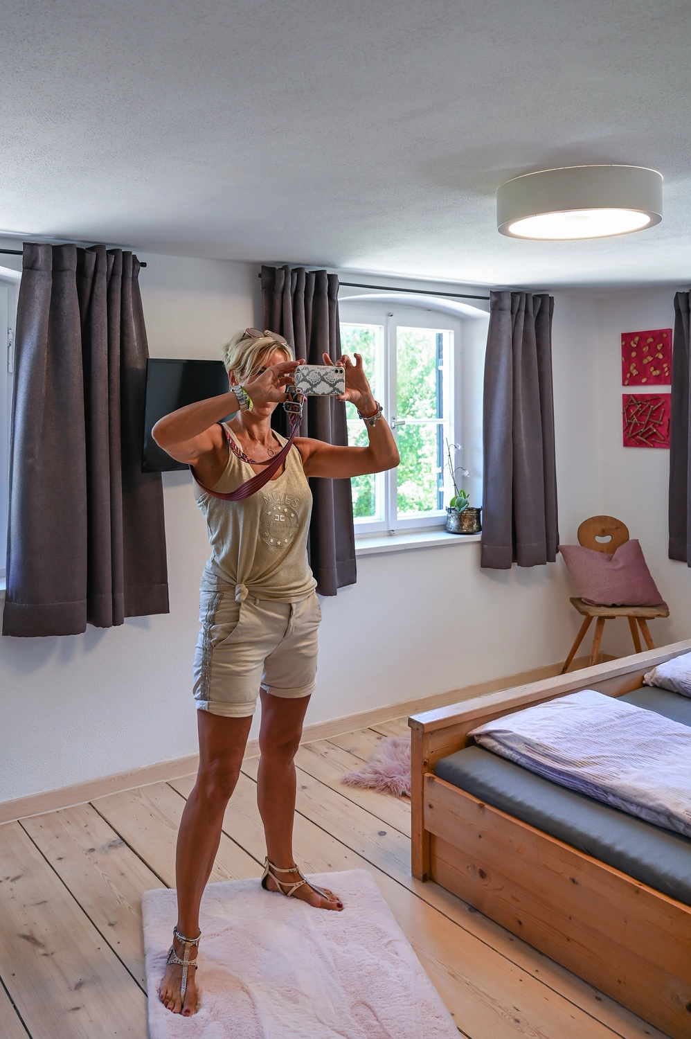 Antjes – Roomstyling – Frau im hellen Sommeroutfit macht im Schlafzimmer ein Selfie mit dem Handy. Holzfußboden, Holzbett und dunkle Vorhänge im Hintergrund.