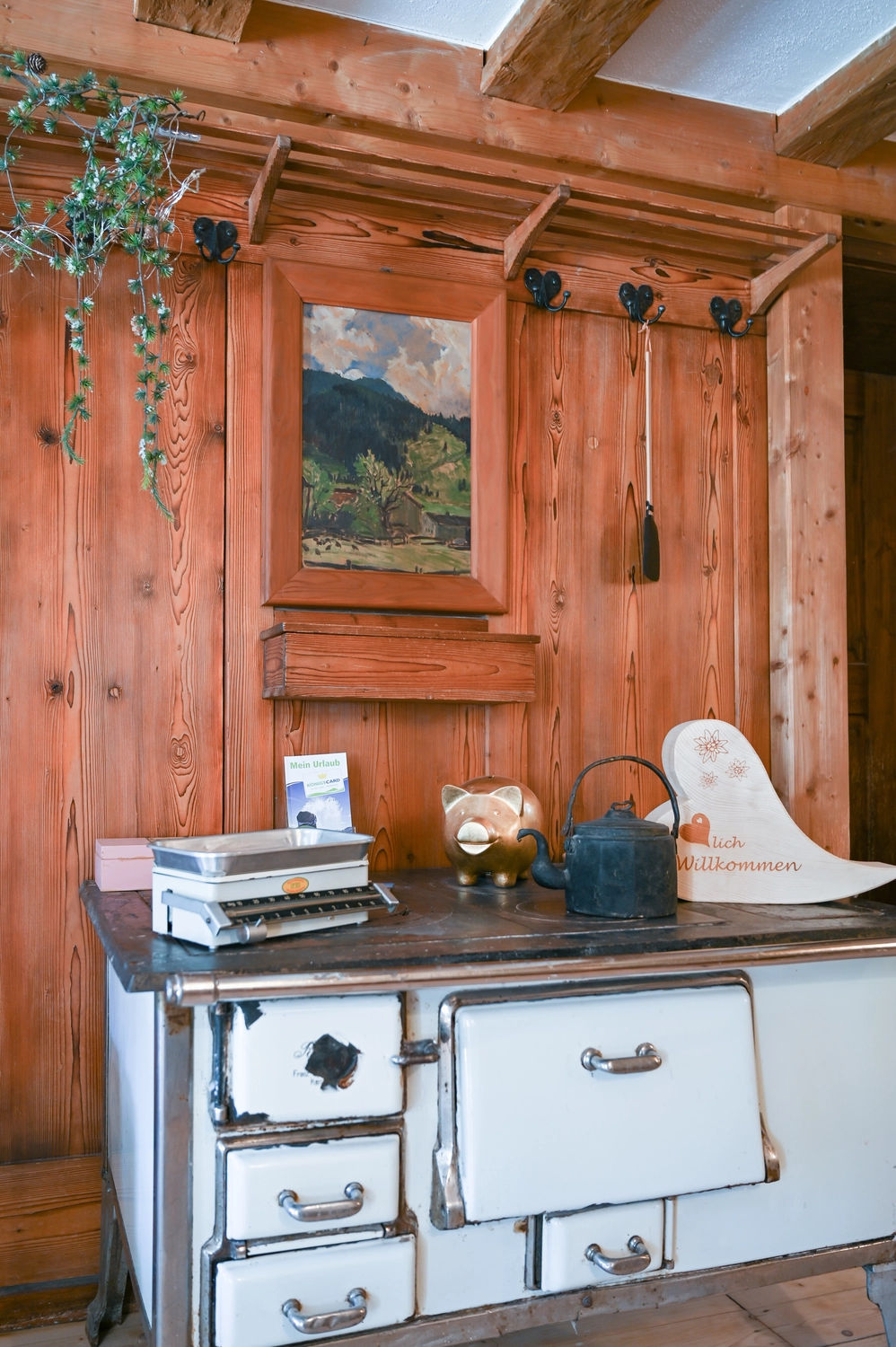 Antjes – Roomstyling –  Eine rustikale Holzstube mit weißer Kommode, darauf Bücher, ein schwarzer Topf und ein Bild. Über der Kommode hängt ein Landschaftsgemälde an einer Holzwand mit Balken und Pflanzen.