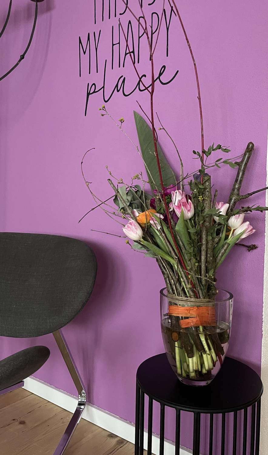 Antjes – Roomstyling –  Eine lila Wand mit dem Schriftzug „This is my happy place“, davor ein schwarzer Beistelltisch mit einer Vase voller bunter Blumen und ein grauer Stuhl daneben.