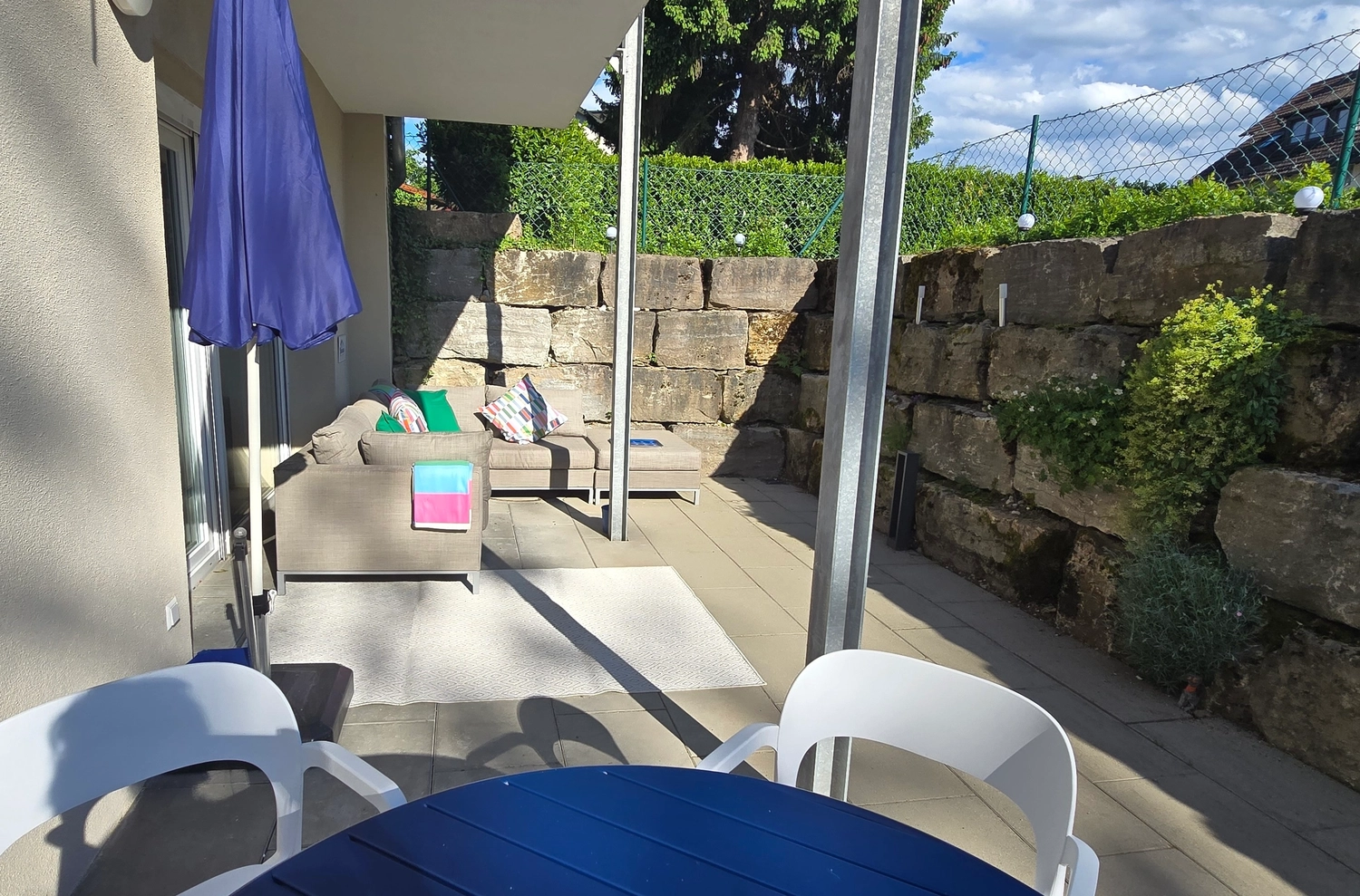 Antjes – Roomstyling – Terrasse mit blauem Tisch, weißen Stühlen und beiger Couch mit bunten Kissen. Dazu ein geschlossener blauer Schirm, Steinmauer und Hecke im Hintergrund.