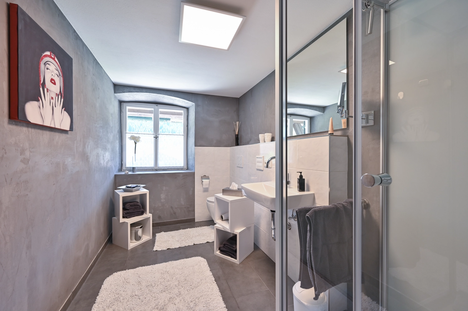 Antjes – Roomstyling –   Nachher: Ein modernes Badezimmer mit grauen Betonwänden, weißem Waschtisch, Glasdusche, Teppichen und minimalistischer Einrichtung.