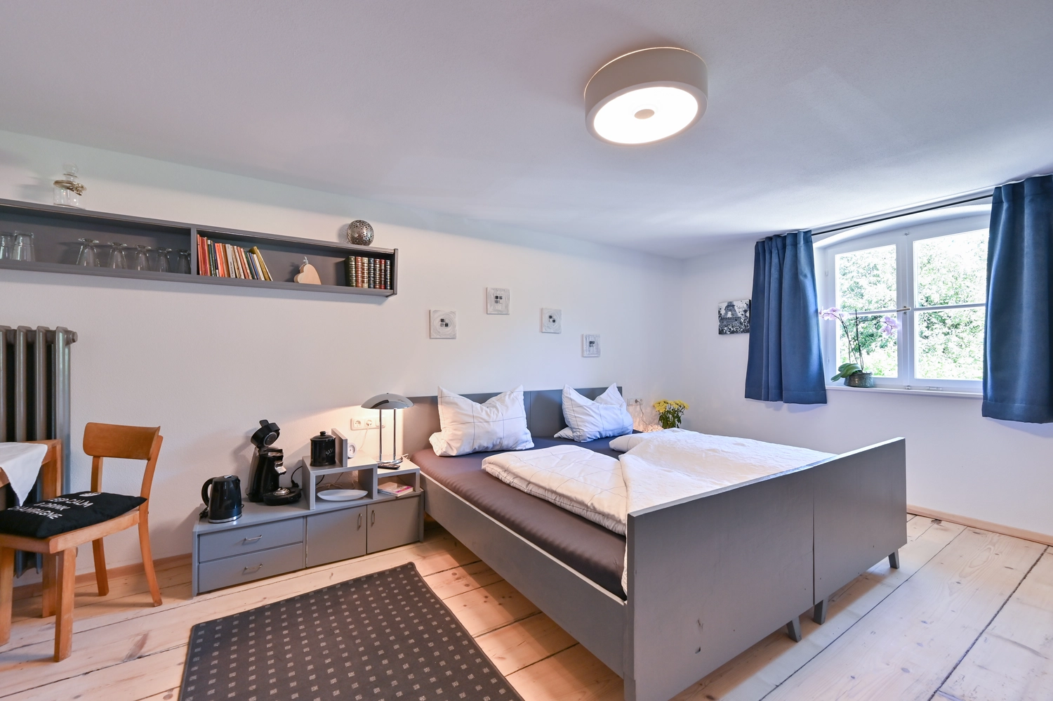 Antjes – Roomstyling –  Nachher: Ein helles, modernisiertes Schlafzimmer mit einem Doppelbett, hellen Holzdielen, blauem Vorhang und minimalistischer Einrichtung.
