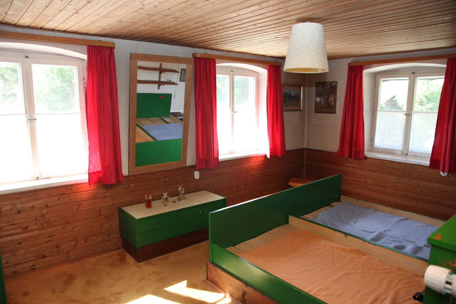 Antjes – Roomstyling – Vorher – Ein rustikales Schlafzimmer mit holzgetäfelten Wänden, leuchtend roten Vorhängen, grünen Möbeln und zwei Einzelbetten unter warmem Sonnenlicht.