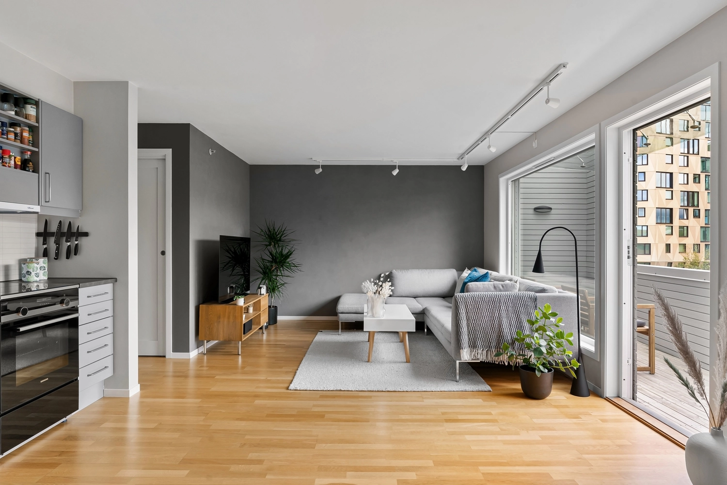 Antjes – Roomstyling – Ein helles, modernes Wohnzimmer mit hellem Holzboden, grauer Akzentwand, Ecksofa, kleinem Couchtisch und großen Fenstern mit Blick nach draußen
