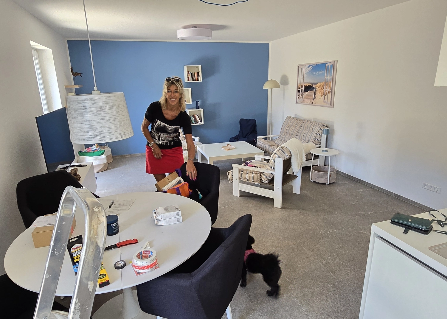 Antjes – Roomstyling – Frau in schwarzem Shirt und rotem Rock steht im Wohnzimmer zwischen Tisch, Sofa und Umzugskartons. Ein kleiner schwarzer Hund ist auch da.