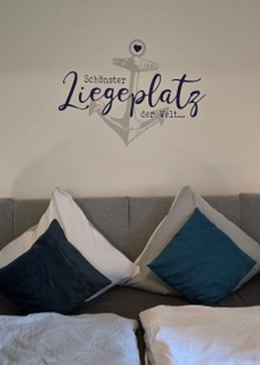 Antjes – Roomstyling –  Ein gemütliches Bett mit grauem Kopfteil, weißen und blauen Kissen, und einem Wandtattoo mit Anker und dem Spruch „Schönster Liegeplatz der Welt...“ darüber.