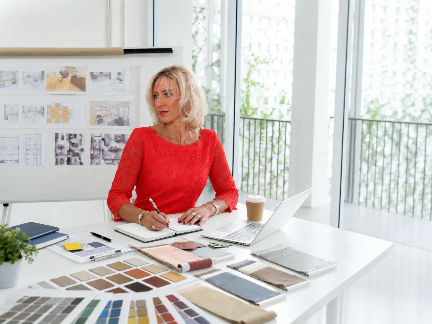 Antjes – Roomstyling – Eine Frau in einem roten Oberteil arbeitet an einem hellen Schreibtisch; hinter ihr sind Designmuster, ein Laptop und Moodboards zu sehen.