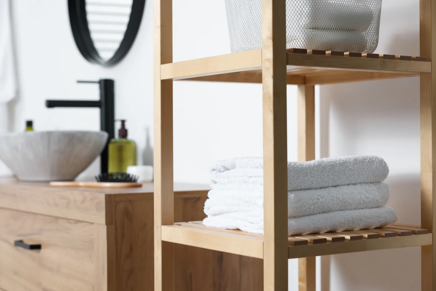 Antjes – Roomstyling –  Ein modernes Badezimmer mit einem Holzregal, auf dem gefaltete weiße Handtücher liegen, neben einem Waschtisch mit Steinwaschbecken und schwarzem Wasserhahn.