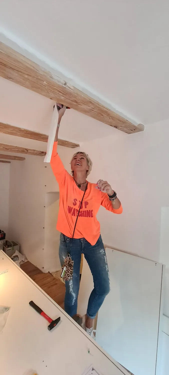 Antjes – Roomstyling –  Person in orange Shirt und Jeans steht auf einer Arbeitsfläche und befestigt einen Holzbalken an der Decke. Werkzeug liegt daneben.