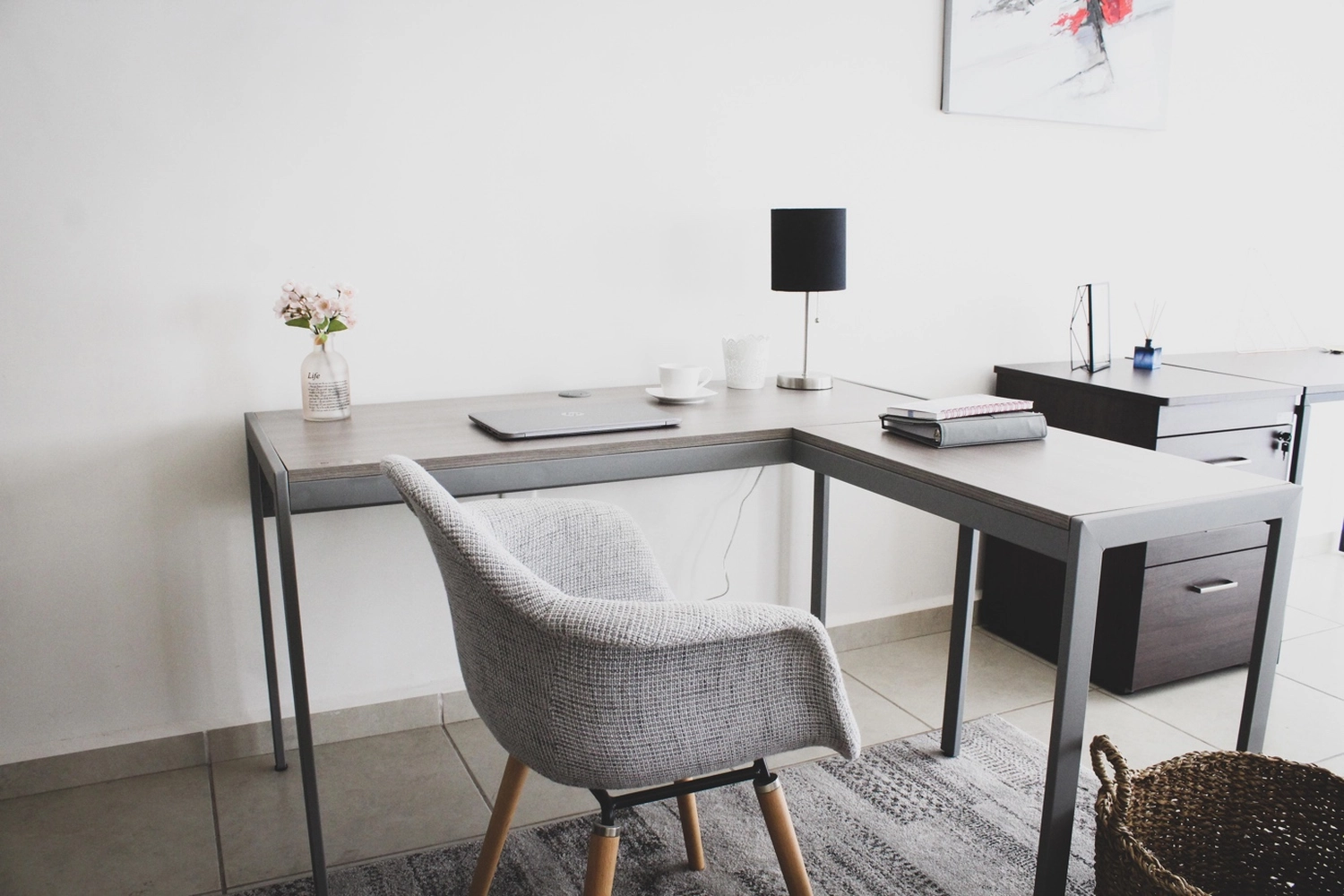 Antjes – Roomstyling – Ein aufgeräumtes, minimalistisches Heimbüro mit einem L-förmigen grauen Schreibtisch, einem grauen Stoffstuhl, einem Laptop, einer Lampe und einer kleinen Blumenvase.