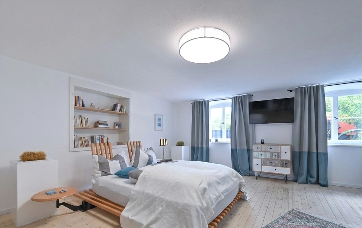 Antjes – Roomstyling –  Ein helles, minimalistisches Schlafzimmer mit Holzbett, weißer Bettwäsche, eingebautem Bücherregal und blaugrauen Vorhängen.