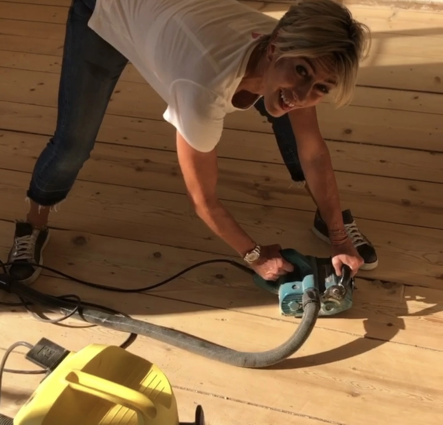 Antjes – Roomstyling – Person im weißen Shirt und Jeans schleift mit einer Maschine einen Holzboden. Ein gelber Staubsauger liegt daneben.