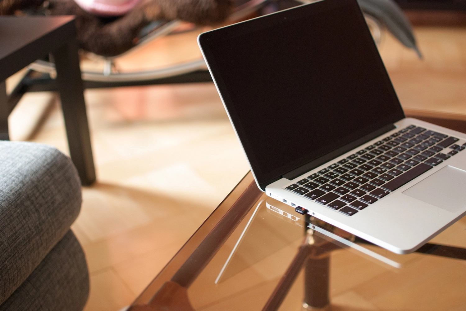Antjes – Roomstyling – Ein silberner Laptop mit schwarzem Bildschirm steht aufgeklappt auf einem gläsernen Couchtisch in einem gemütlichen Wohnzimmer.